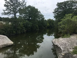 Onion Creek. In the foreground, rocky ledges hang over both sides of the still creek. Beyond the rocky outcroppings, the water reflects the dense lines of trees on both sides.
